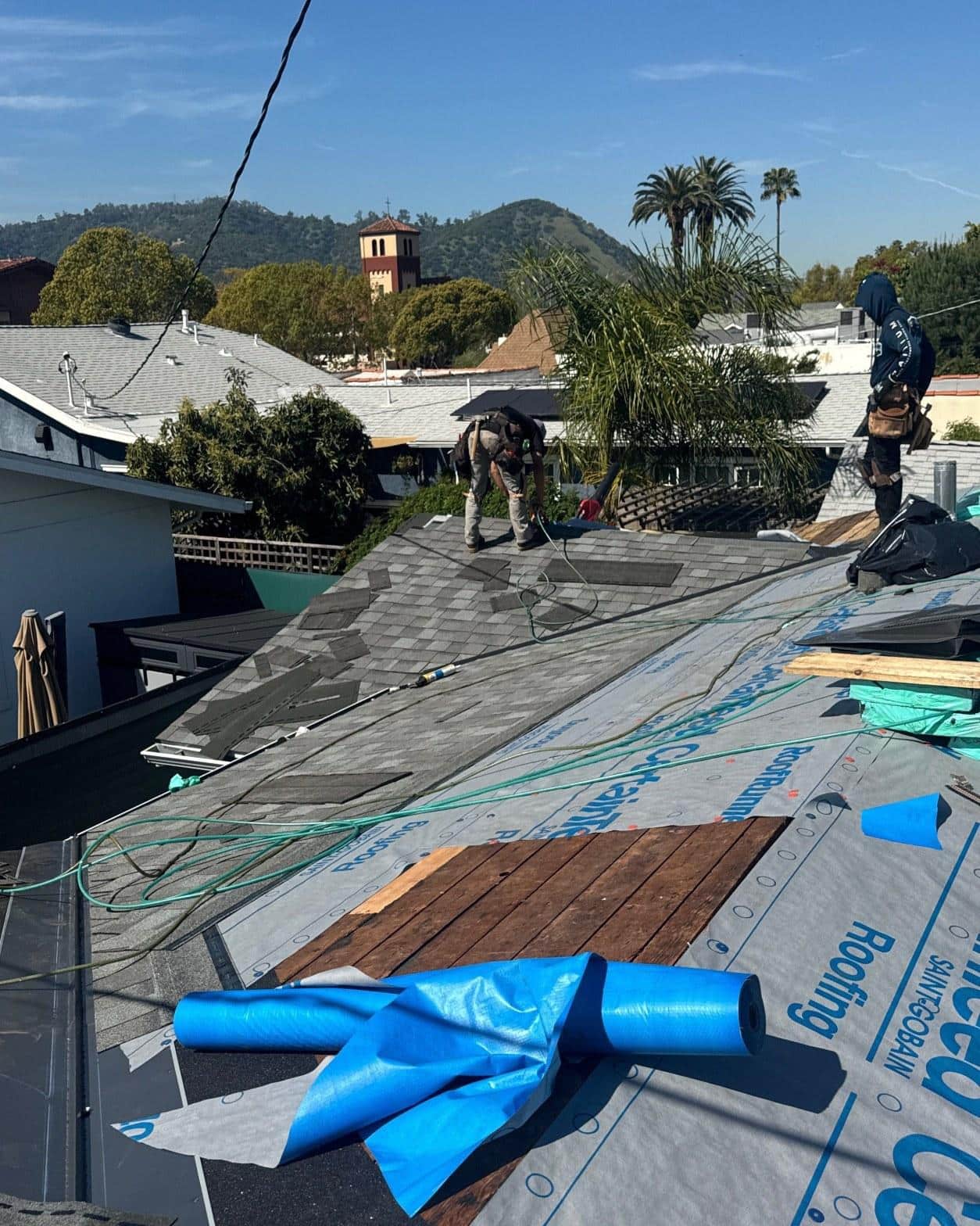 Two roofers laying gray asphalt shingles on residential home with underlayment and blue tarp visible, hills in background.