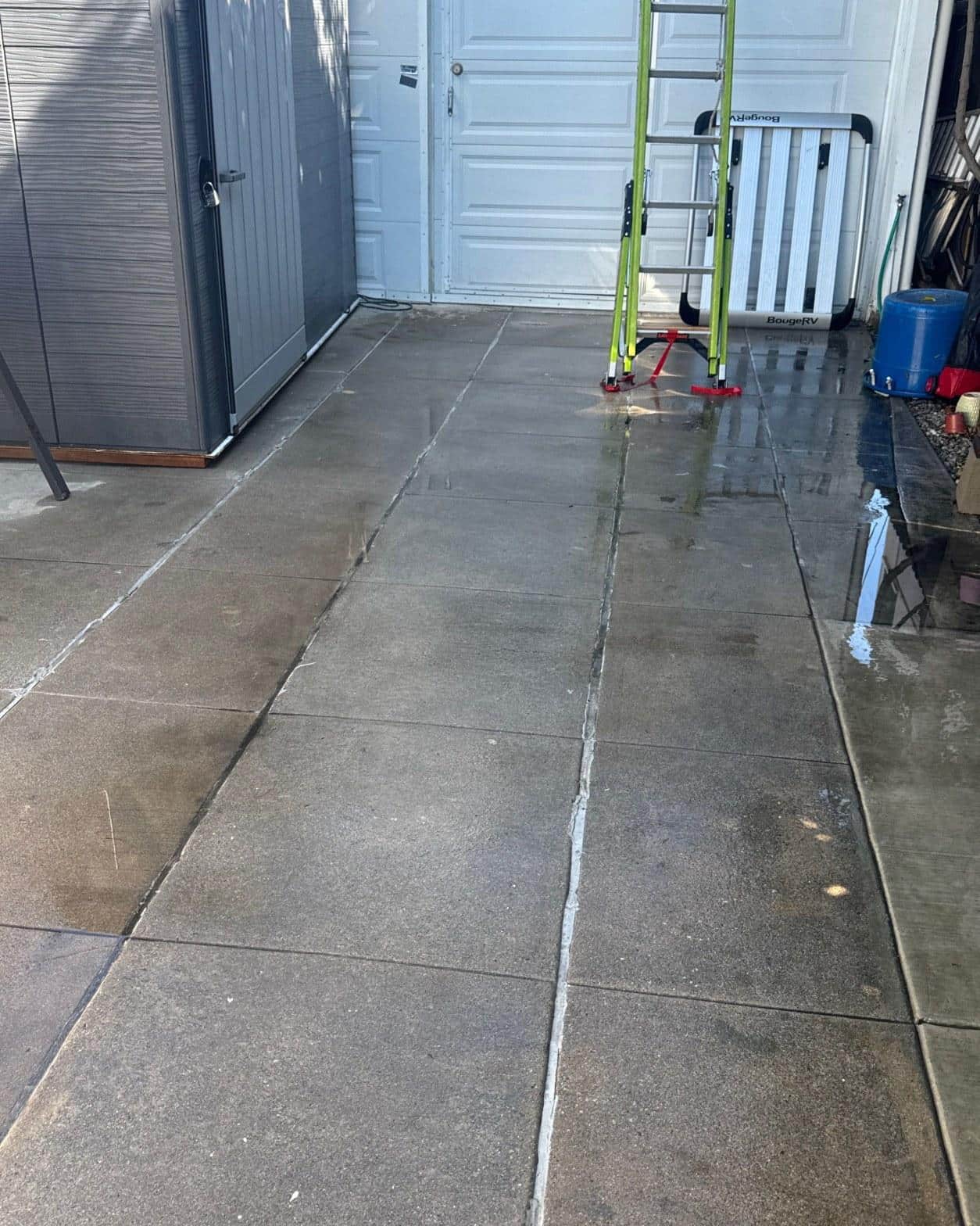 Freshly pressure-washed concrete driveway with expansion joints, green ladder, and gray storage shed near garage.