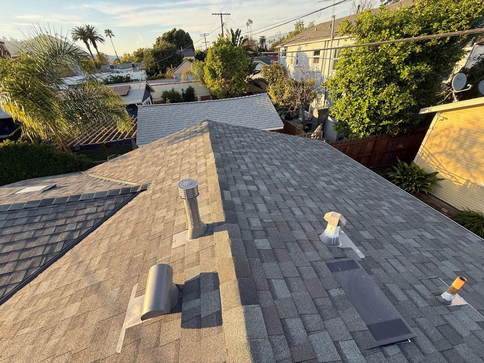 Newly installed gray asphalt shingle roof with plumbing and exhaust vents, residential neighborhood background.