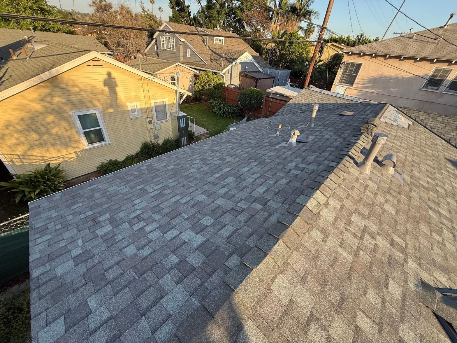 Freshly installed gray asphalt shingle roof with plumbing pipe vents, suburban neighborhood homes visible in background.