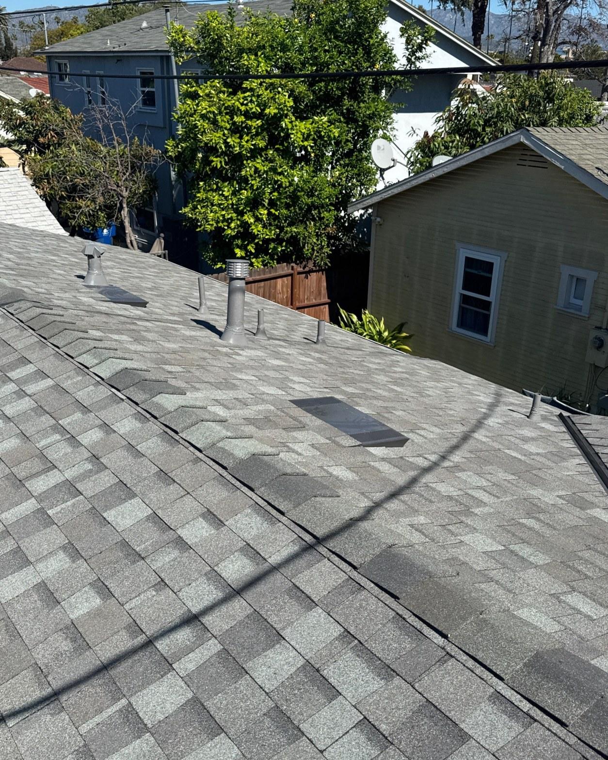 Newly installed gray asphalt shingle roof with metal pipe vents, valley flashing, and residential neighborhood in background.