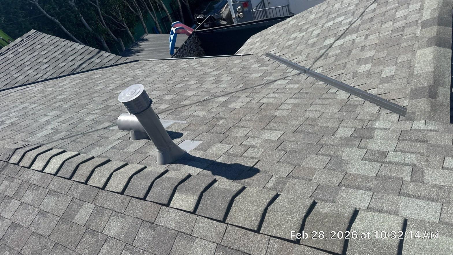 Gray PVC vent pipe with cap installed through asphalt shingle roof with ridge vent visible nearby.