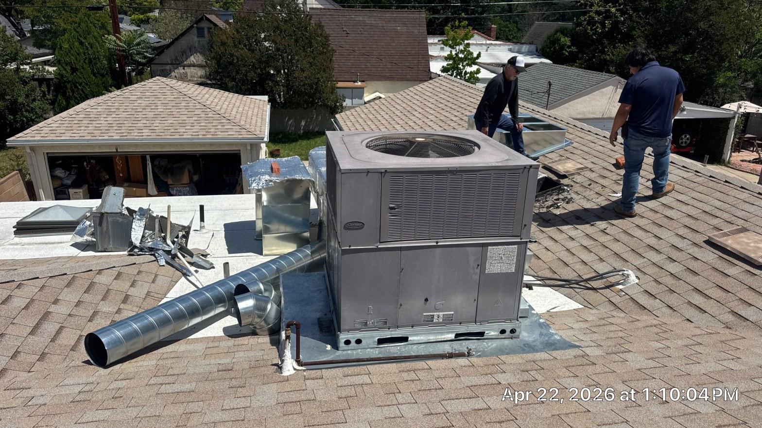 Two HVAC technicians installing a Carrier rooftop package unit with ductwork on a residential shingled roof.