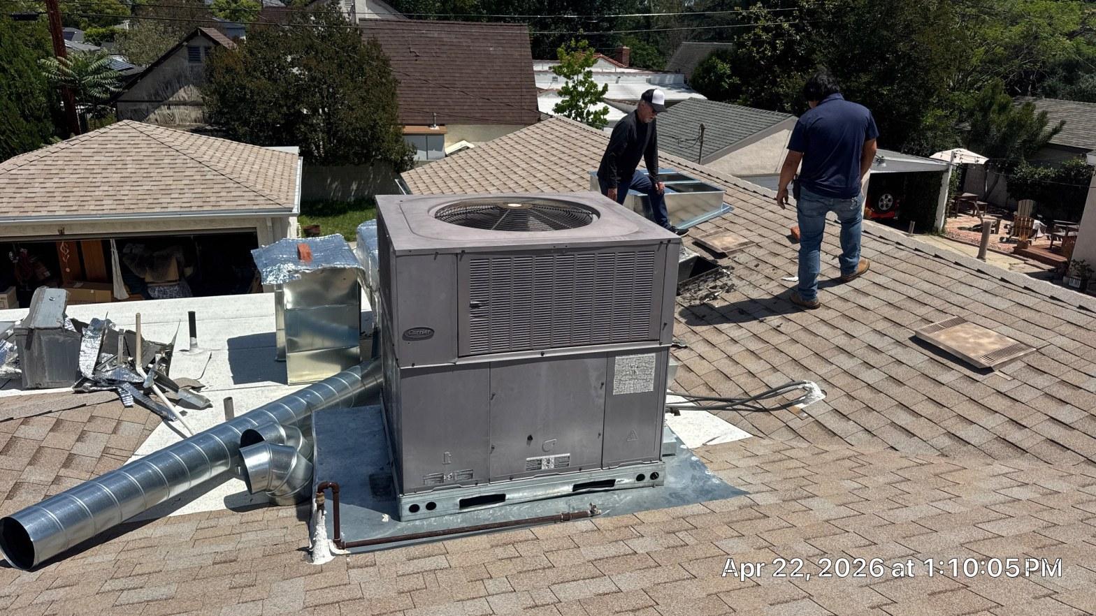 carrier-rooftop-package-unit-installation-burbank-ca-820 Two HVAC technicians installing a Carrier rooftop packaged AC unit with ductwork on asphalt shingle roof.