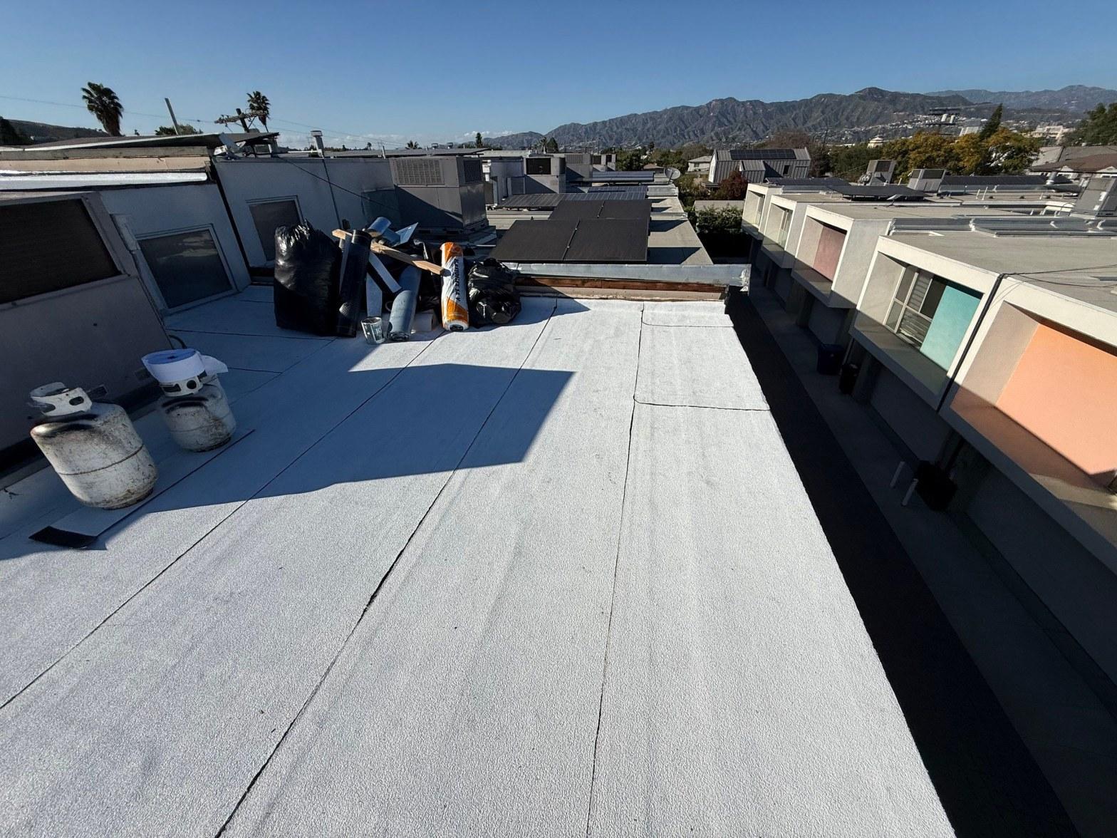 foam-roof-coating-flat-commercial-roof-los-angeles-perlita-a-1001 White elastomeric flat roof coating on urban building with roofing debris, tools, and San Gabriel Mountains in background.
