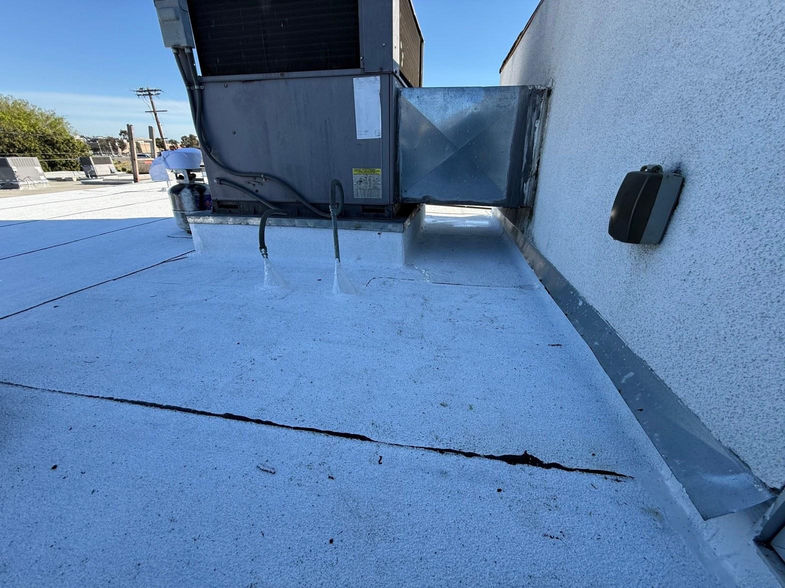 foam-roof-coating-flat-commercial-roof-los-angeles-perlita-a-1003 Commercial flat roof with large crack in white coating, standing water, and rooftop HVAC package unit on sunny day.