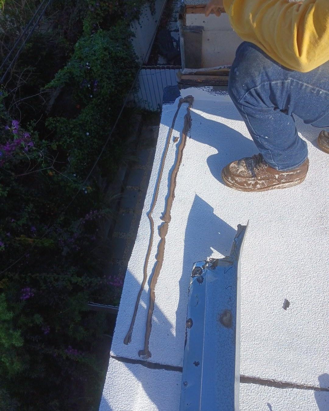 foam-roof-coating-flat-commercial-roof-los-angeles-perlita-a-1005 Worker in yellow shirt and jeans sealing a crack on a white flat roof surface with brown sealant caulk.