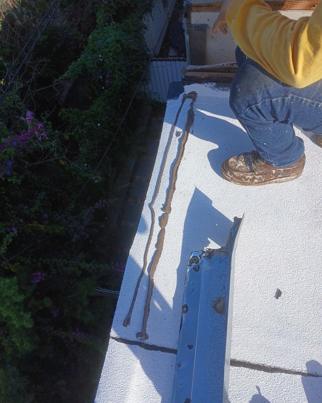 foam-roof-coating-flat-commercial-roof-los-angeles-perlita-a-1007 Roofer in yellow shirt and jeans sealing cracks on white flat roof with brown sealant bead.