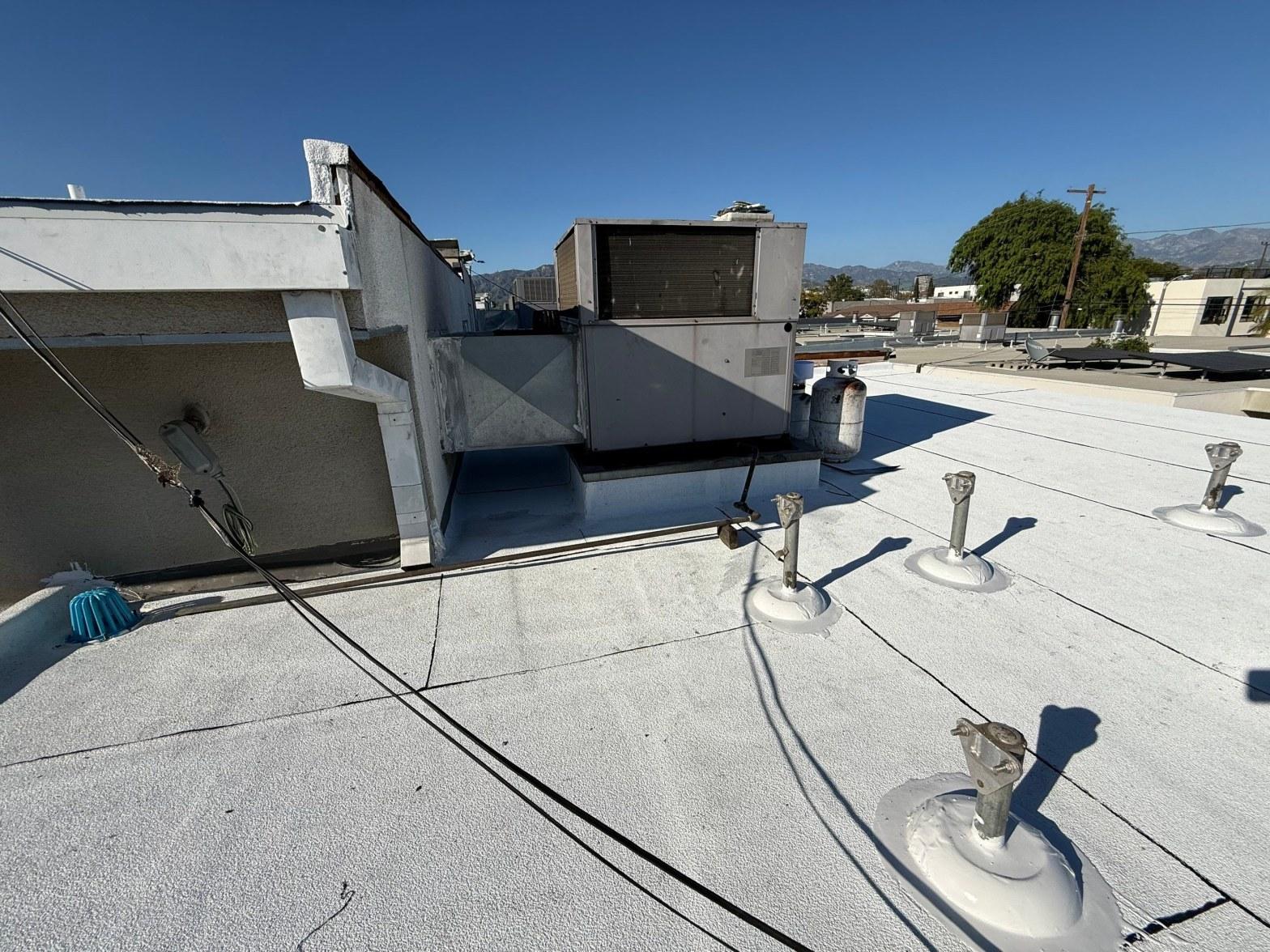 foam-roof-coating-flat-commercial-roof-los-angeles-perlita-a-1009 Commercial flat roof with elastomeric coating, plumbing pipe boots, rooftop HVAC unit, and propane tank, mountains in background.