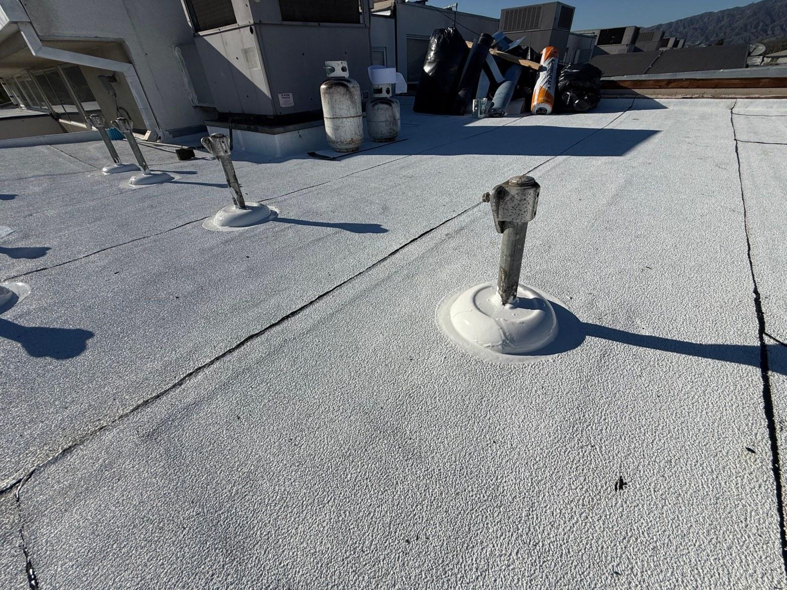 foam-roof-coating-flat-commercial-roof-los-angeles-perlita-a-1011 Commercial flat roof showing pipe penetrations with white sealant boots, surface cracks, and rooftop HVAC equipment in background.
