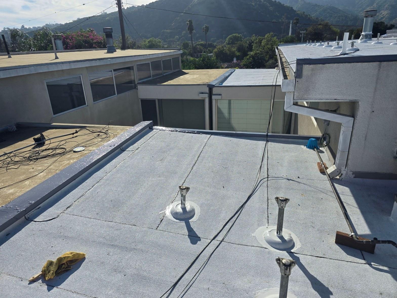 foam-roof-coating-flat-commercial-roof-los-angeles-perlita-a-982 Flat commercial roof with gray membrane, three metal vent pipes, and wiring, mountains visible in background.