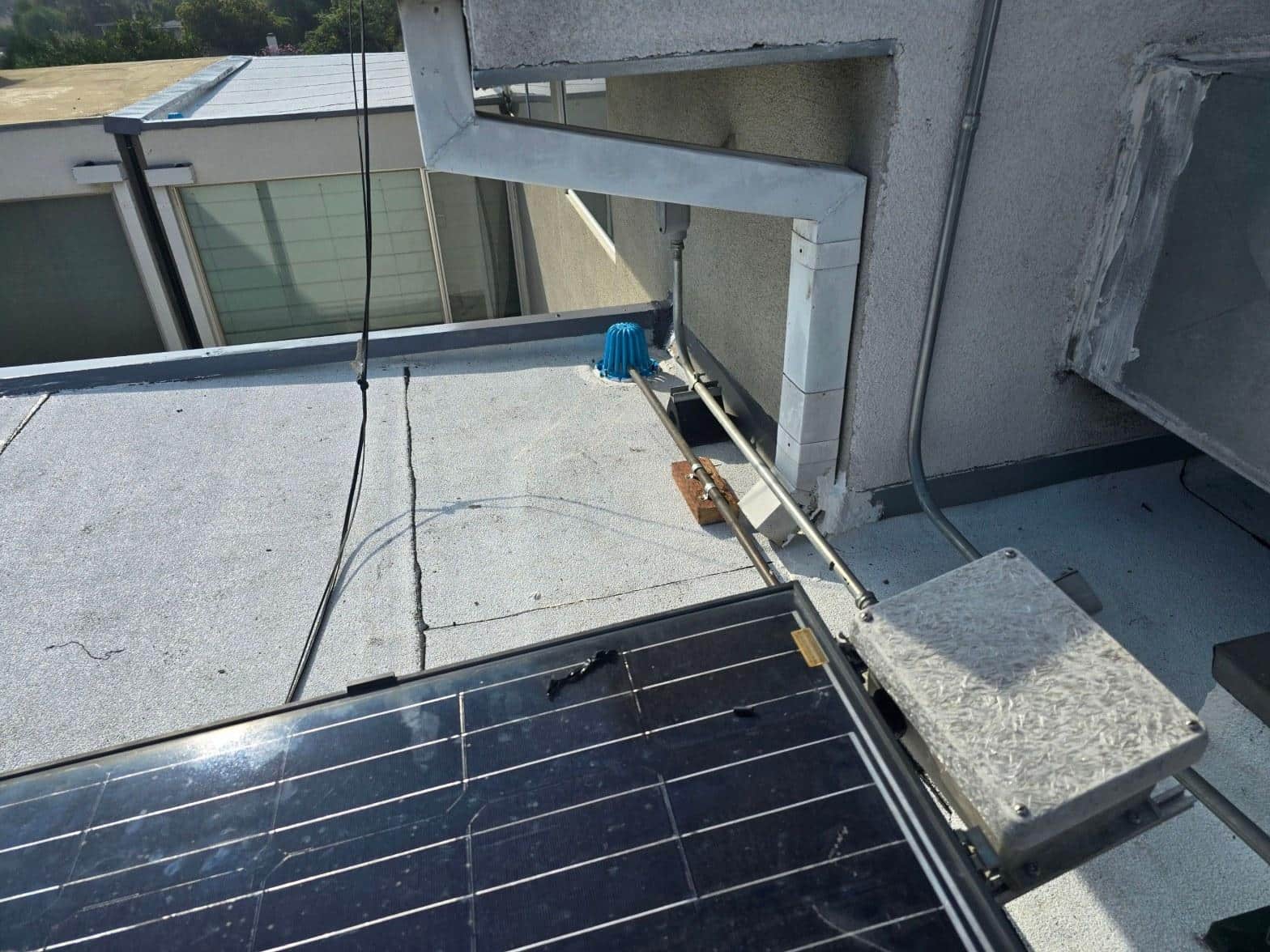 foam-roof-coating-flat-commercial-roof-los-angeles-perlita-a-988 Solar panels with junction box, loose wiring, and blue drain cap on flat gravel roof near stucco wall.