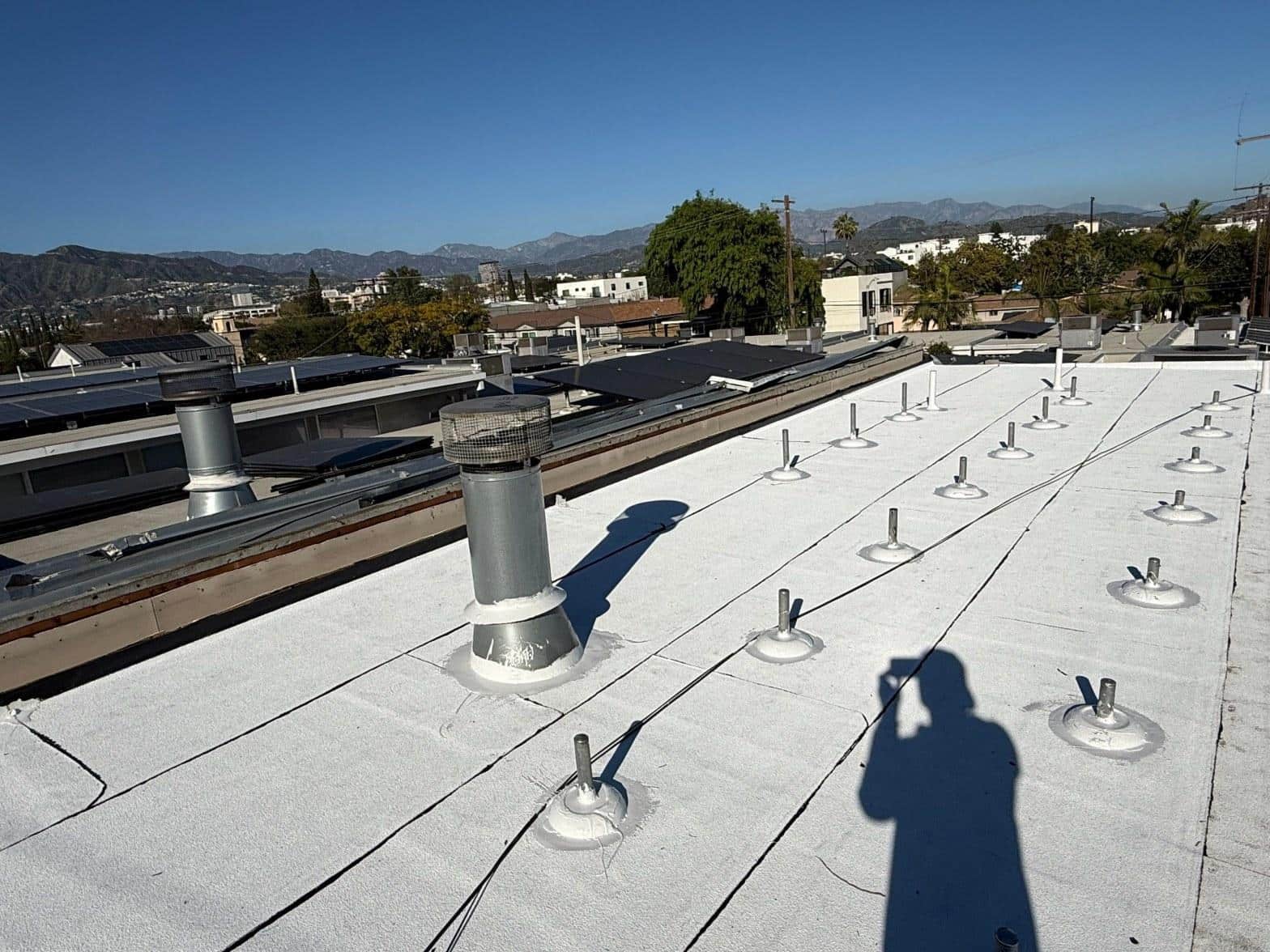 foam-roof-coating-flat-commercial-roof-los-angeles-perlita-a-995 White-coated commercial flat roof with pipe boots, vent stacks, and city skyline with mountains in background.