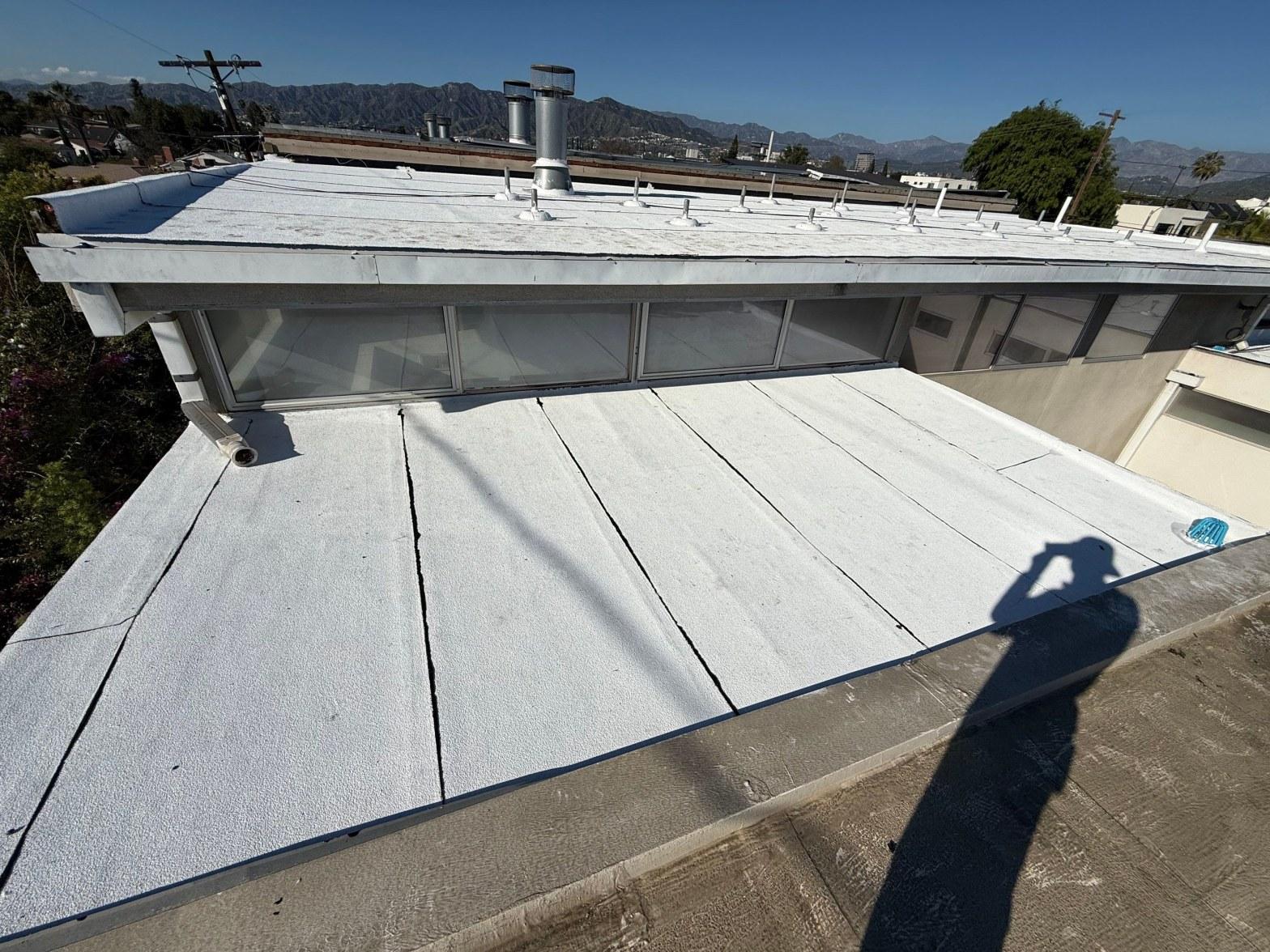 foam-roof-coating-flat-commercial-roof-los-angeles-perlita-a-996 White spray foam flat roof with surface cracks, vent pipes, and clerestory windows on commercial building, mountains in background.