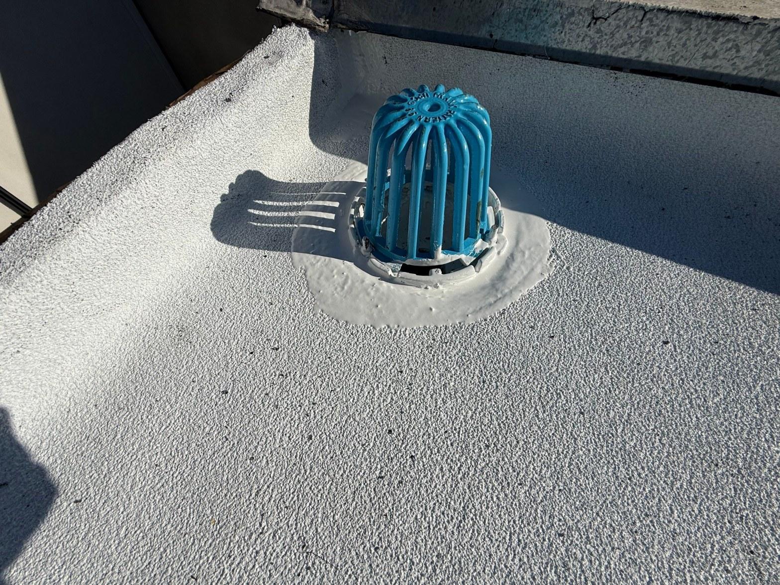 foam-roof-coating-flat-commercial-roof-los-angeles-perlita-a-997 Blue cast iron dome strainer roof drain sealed with elastomeric coating on white flat roof surface.