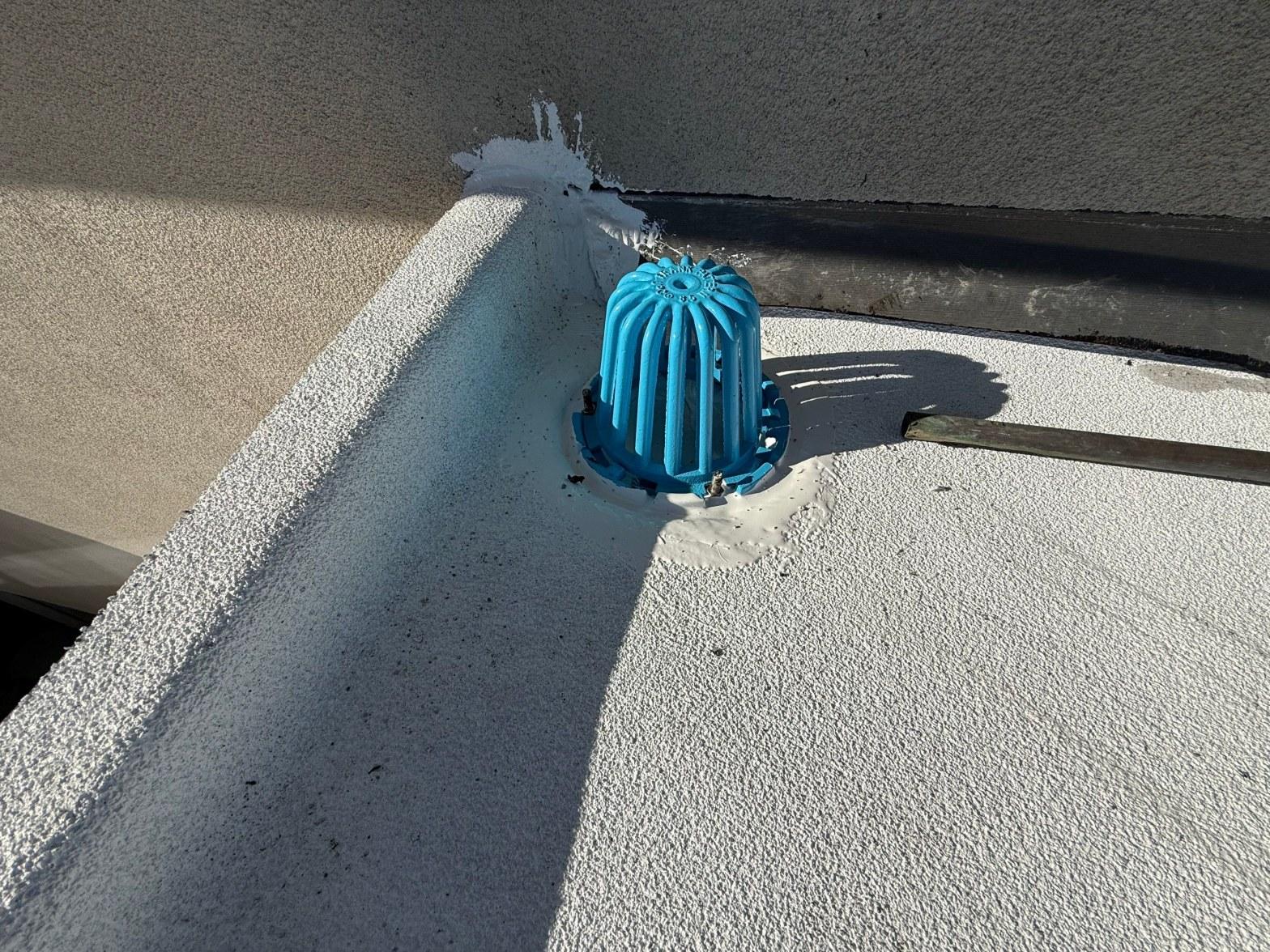 foam-roof-coating-flat-commercial-roof-los-angeles-perlita-a-998 Blue dome-shaped roof drain strainer cover installed in corner of flat white elastomeric coated roof near parapet wall.