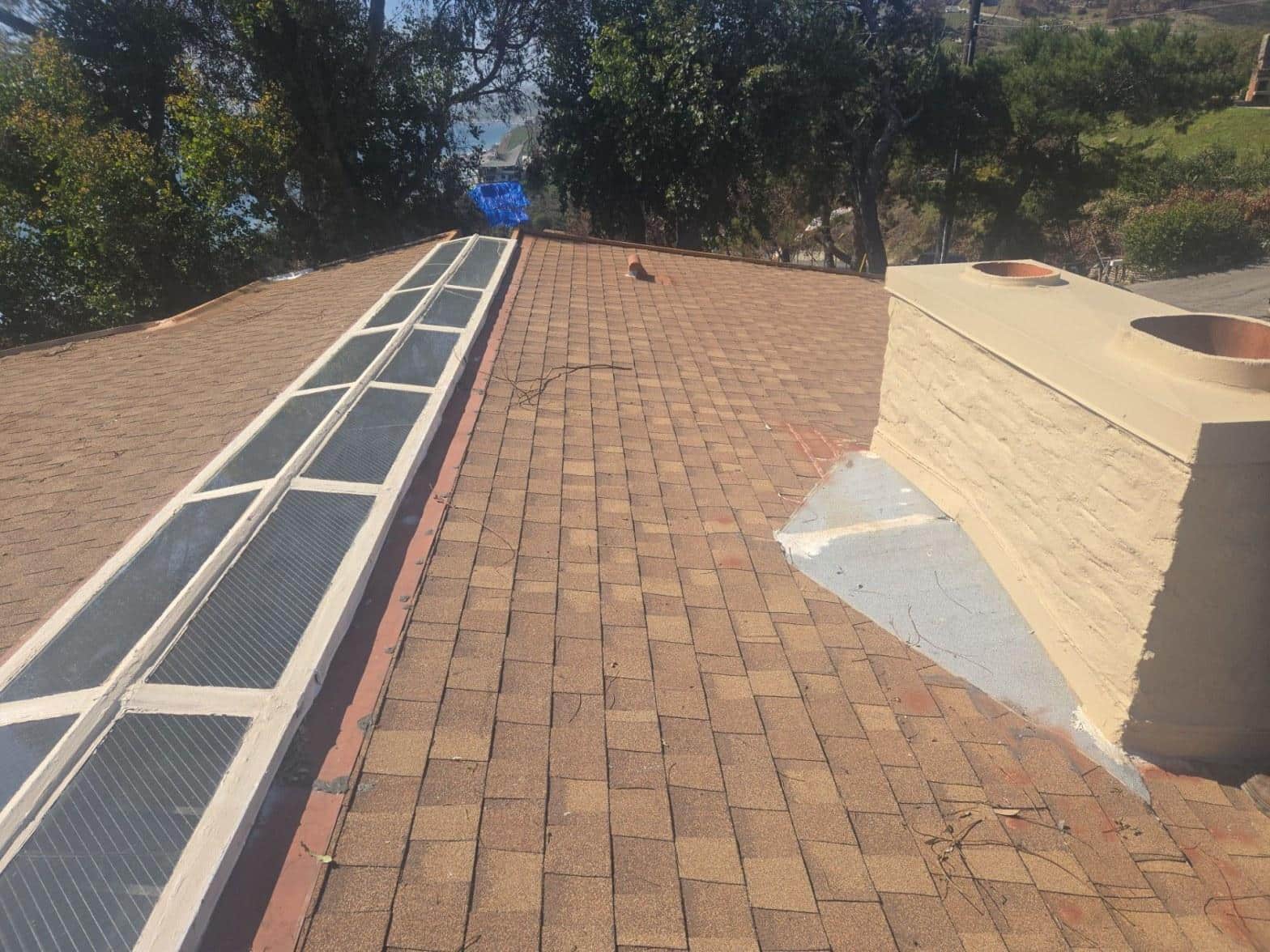 Brown asphalt shingle roof with long skylight, white stucco chimney, and exposed white flashing patch near chimney base.