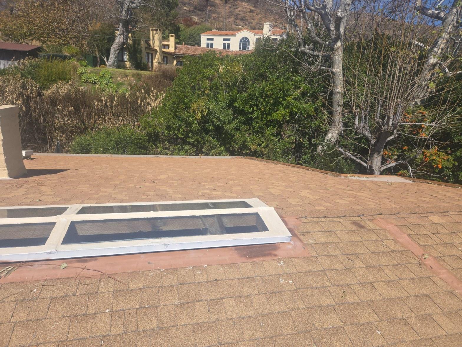 Removed skylight frame with mesh screens lying on residential asphalt shingle roof, hillside homes in background.