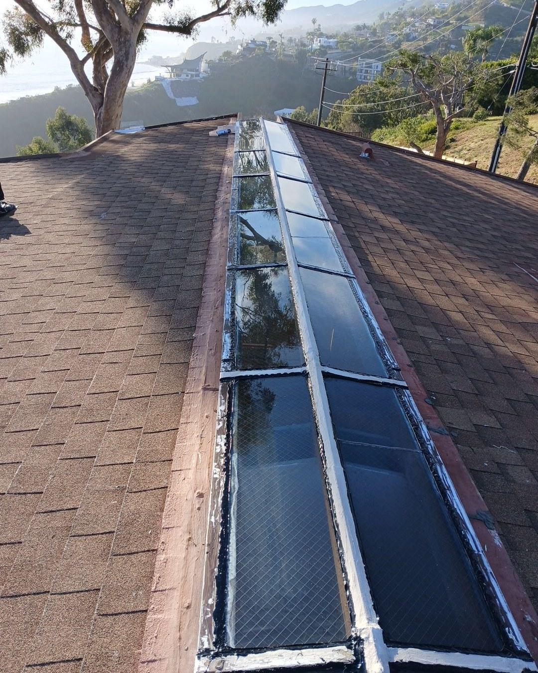 Series of glass skylights sealed with white caulk along roof ridge, asphalt shingles, coastal hillside view.