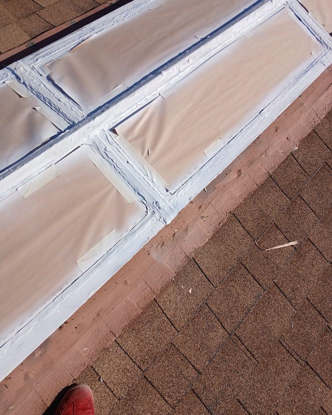 Close-up of rooftop skylight frame with fresh white caulk sealant on brown asphalt shingle roof.