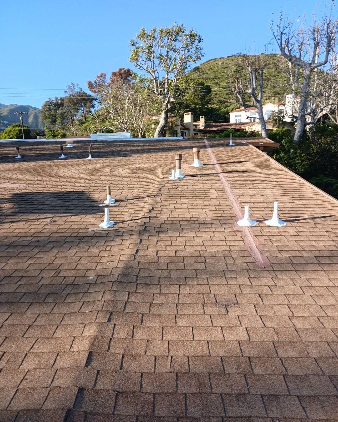 Brown asphalt shingle roof with several white and metal plumbing vent pipes, hillside neighborhood in background.