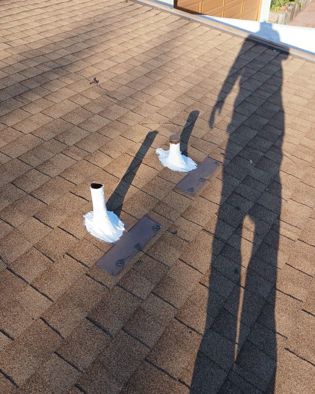 Two white pipe penetrations with caulk sealant on metal flashing plates over brown asphalt shingle roof.