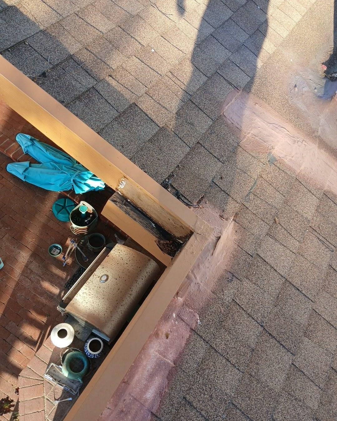 Severely rotted wood fascia board visible at roof edge with asphalt shingles and patio area below.