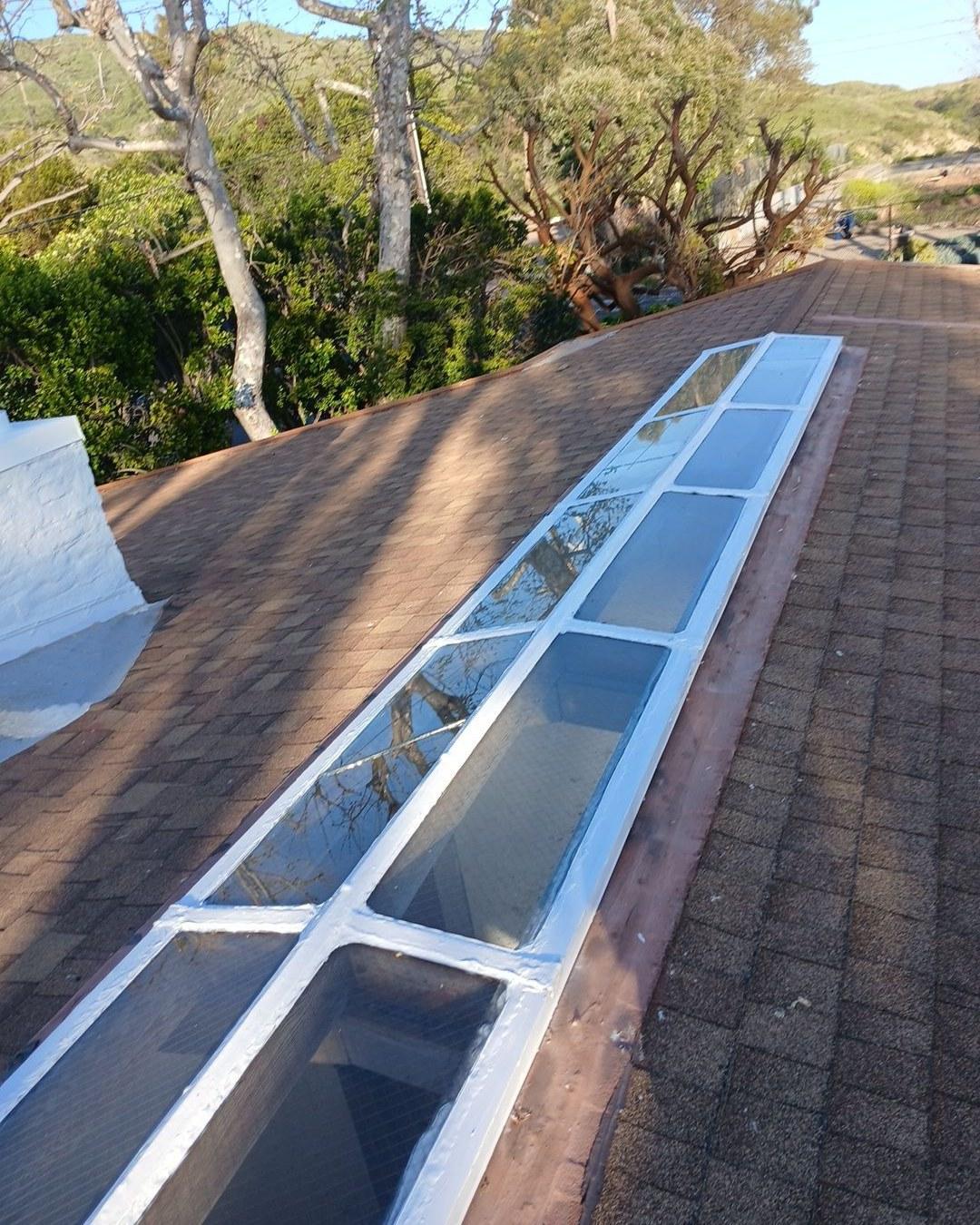 Long narrow multi-panel glass skylight with white frame set into brown asphalt shingle roof on hillside home.