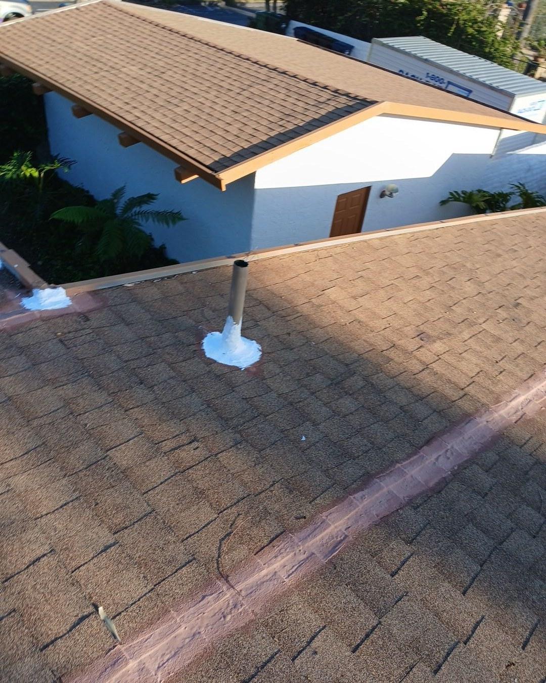Metal pipe boot penetration with white sealant caulk on brown asphalt shingle roof, neighboring house in background.