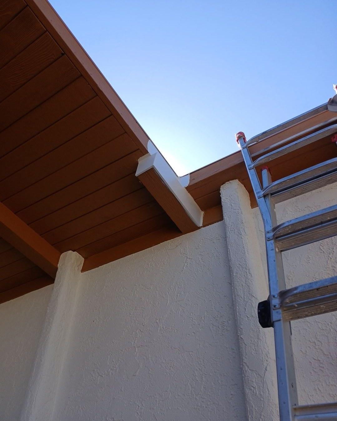 Aluminum ladder leaning against white stucco exterior wall below brown wood soffit and white fascia corner.