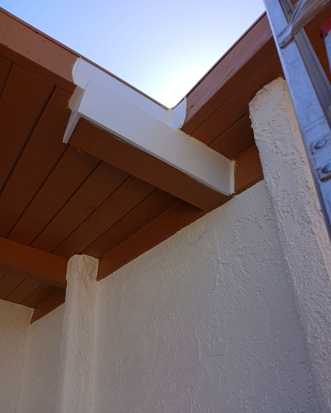White vinyl rain gutter mitered corner joint mounted under brown painted wood soffit on stucco exterior wall.