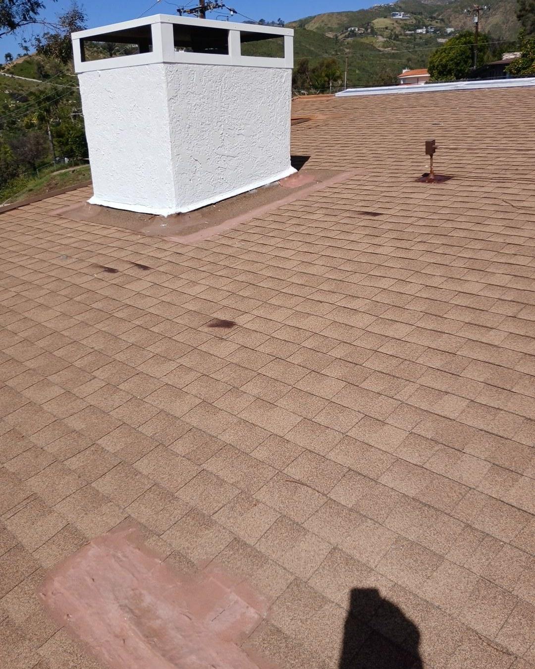 Newly installed tan asphalt shingle roof with white stucco chimney and roof vent, hillside background.
