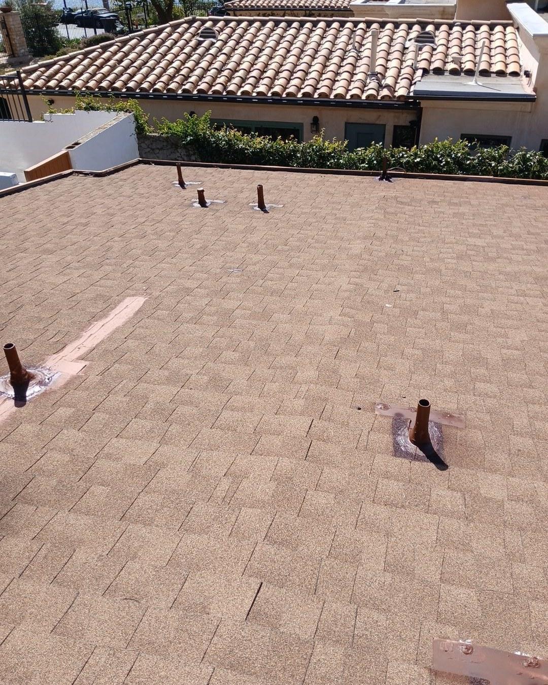 Newly installed brown asphalt shingle roof with plumbing vent pipes, adjacent to Spanish tile roof building.