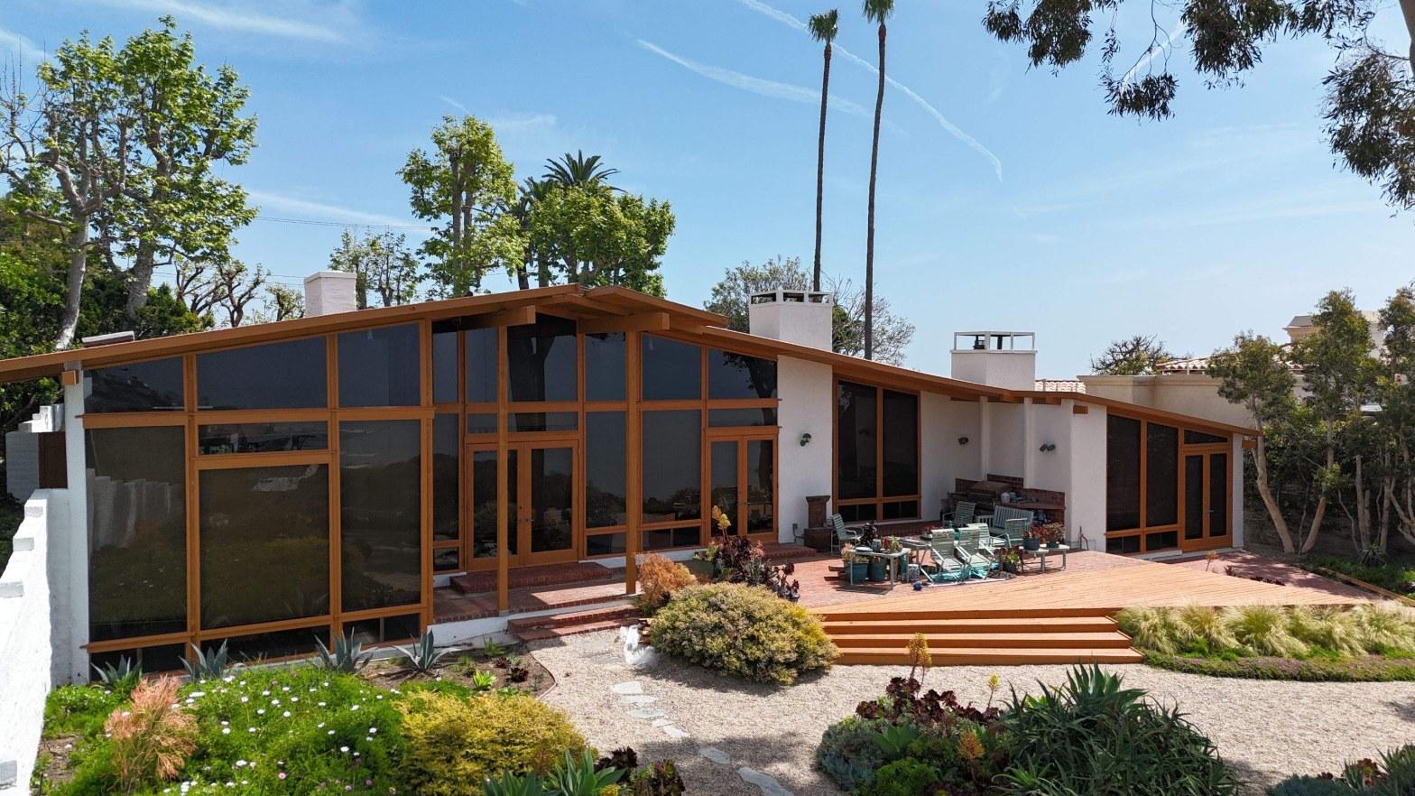 Mid-century modern single-story house with large wood-framed glass windows, outdoor deck, and desert landscaping.