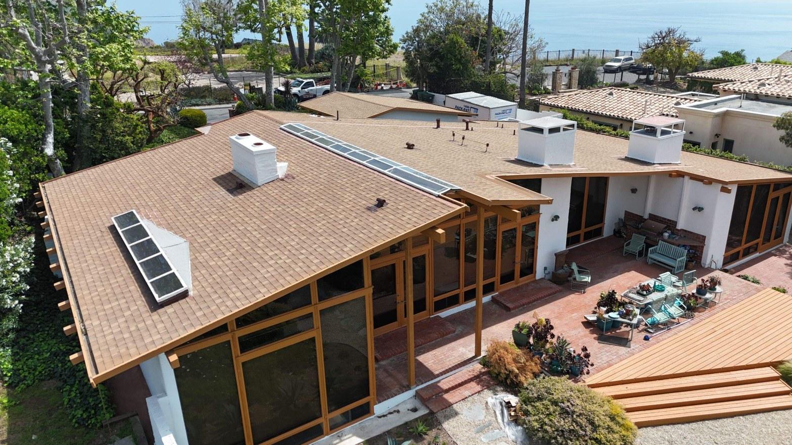 Aerial drone photo of tan asphalt shingle roof with solar panels, white chimneys, and courtyard patio on coastal home.