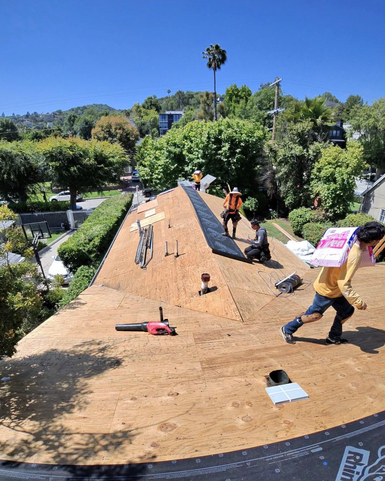 shingle-reroof-14819-sutton-st-los-angeles-557 Three workers laying black roofing underlayment on bare plywood roof deck, suburban California neighborhood in background.