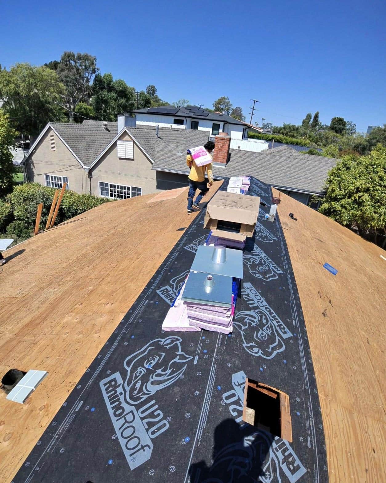 shingle-reroof-14819-sutton-st-los-angeles-558 Roofer carrying shingle bundle on plywood roof deck with RhinoRoof U20 underlayment, suburban neighborhood background.