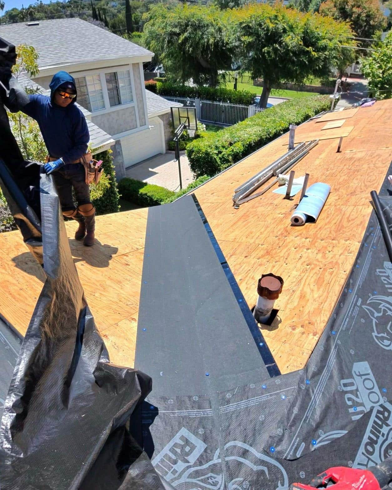shingle-reroof-14819-sutton-st-los-angeles-562 Roofer in blue hoodie rolling out RhinoRoof underlayment on exposed plywood sheathing during roof replacement.