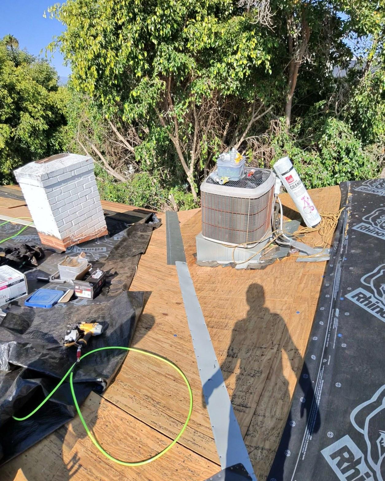 shingle-reroof-14819-sutton-st-los-angeles-565 Residential roof replacement in progress showing plywood decking, rooftop AC condenser unit, brick chimney, and RhinoRoof underlayment partially installed.