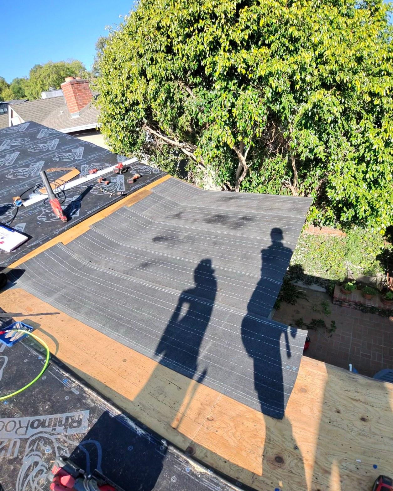 shingle-reroof-14819-sutton-st-los-angeles-566 RhinoRoof underlayment being installed on plywood roof deck with nail gun and tools visible, two shadows of workers cast on surface.