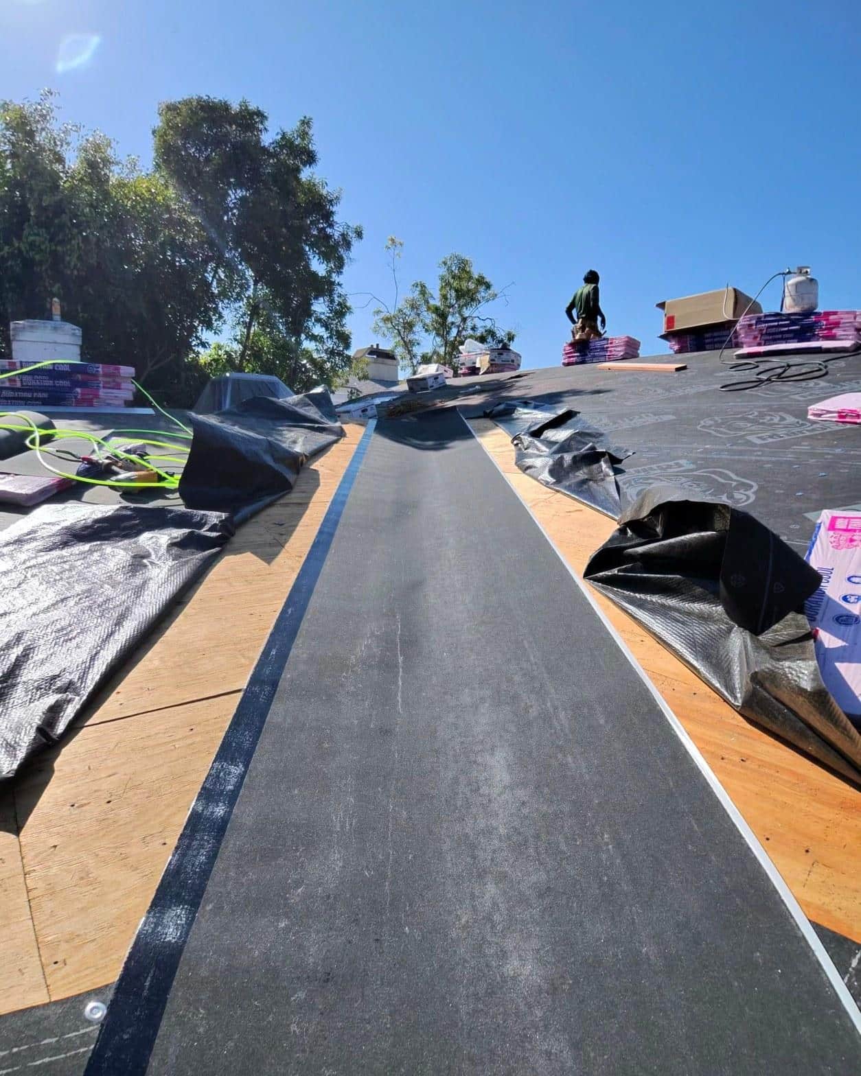 shingle-reroof-14819-sutton-st-los-angeles-568 Roofing contractor standing on steep plywood roof deck with black underlayment rolls and shingle bundles on sunny day.