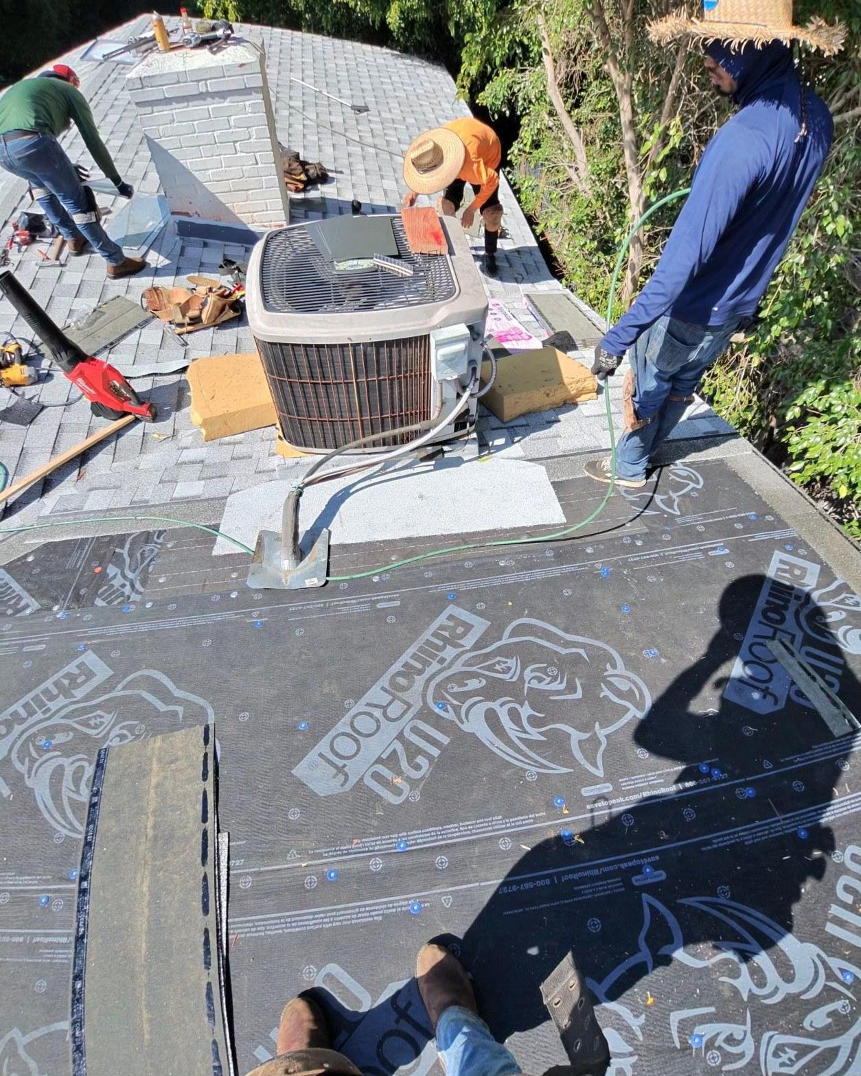 shingle-reroof-14819-sutton-st-los-angeles-571 Three roofers laying RhinoRoof U20 underlayment on flat roof section beside HVAC condenser unit outdoors.