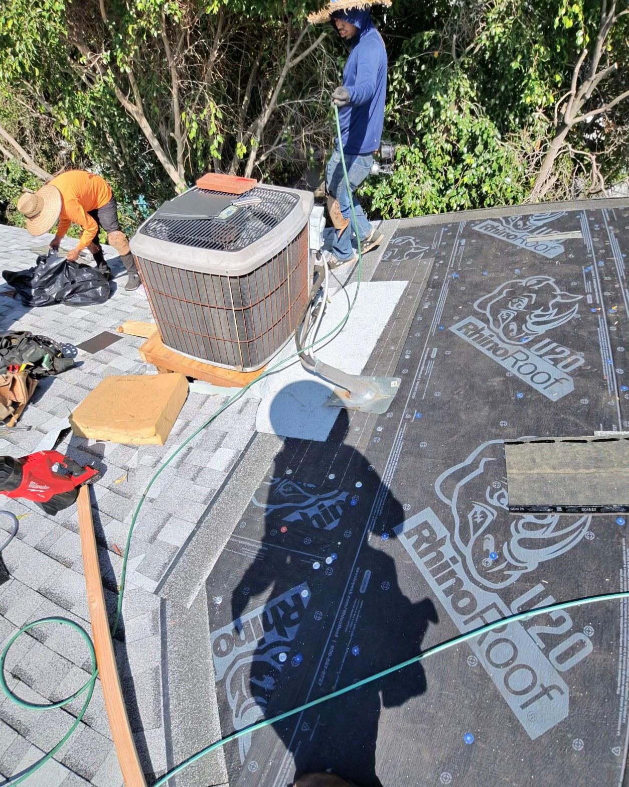 shingle-reroof-14819-sutton-st-los-angeles-572 Two workers laying RhinoRoof U20 synthetic underlayment on flat roof section next to HVAC condenser unit.