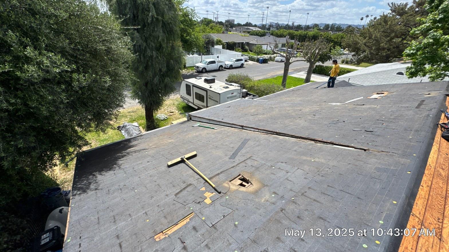 Worn asphalt shingle roof during tear-off showing hole in decking, worker standing at ridge, residential neighborhood.