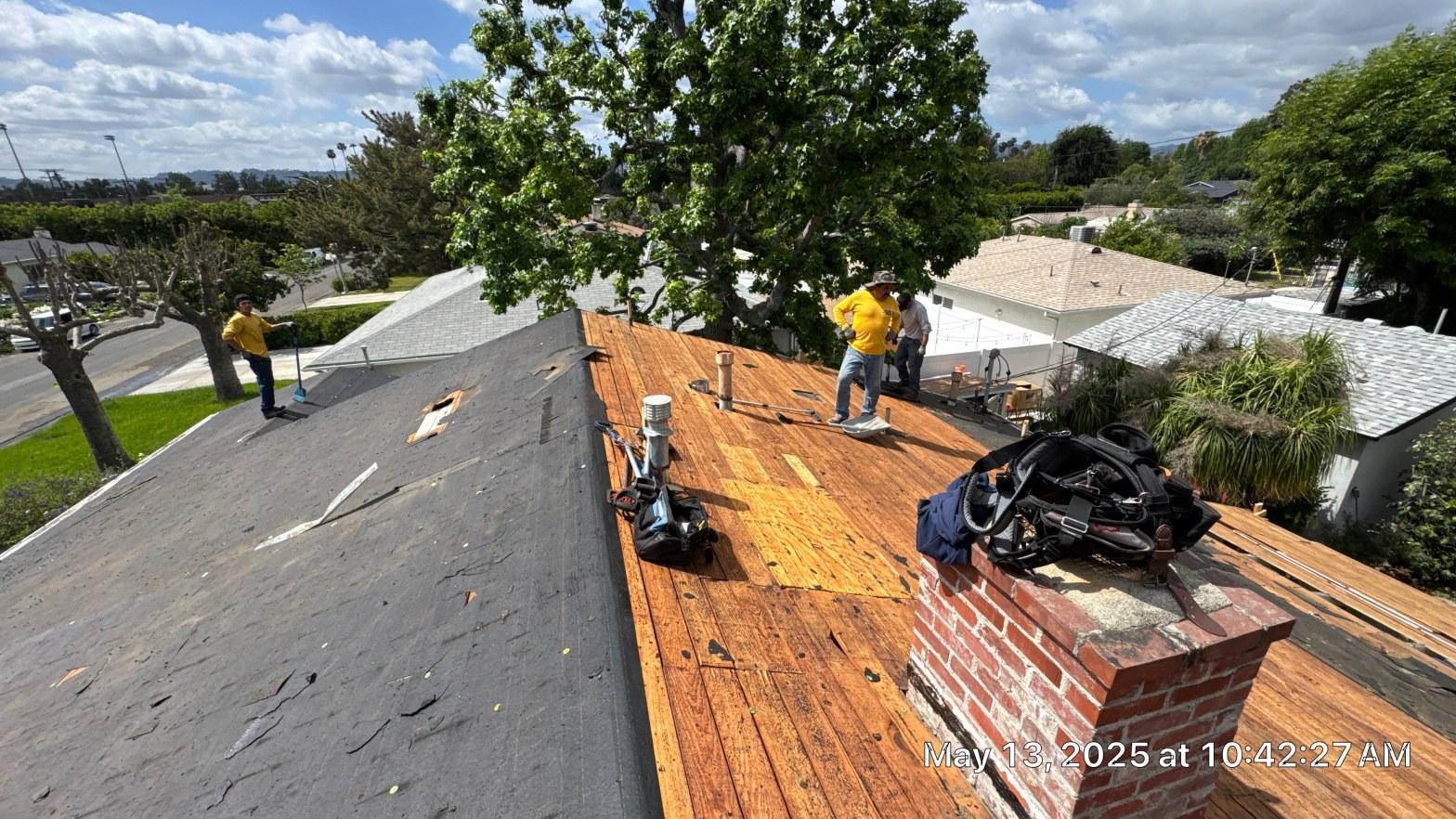 shingle-reroof-6643-saloma-ave-los-angeles-664 Roofing crew in yellow shirts tearing off asphalt shingles, exposing wood board decking with brick chimney visible.