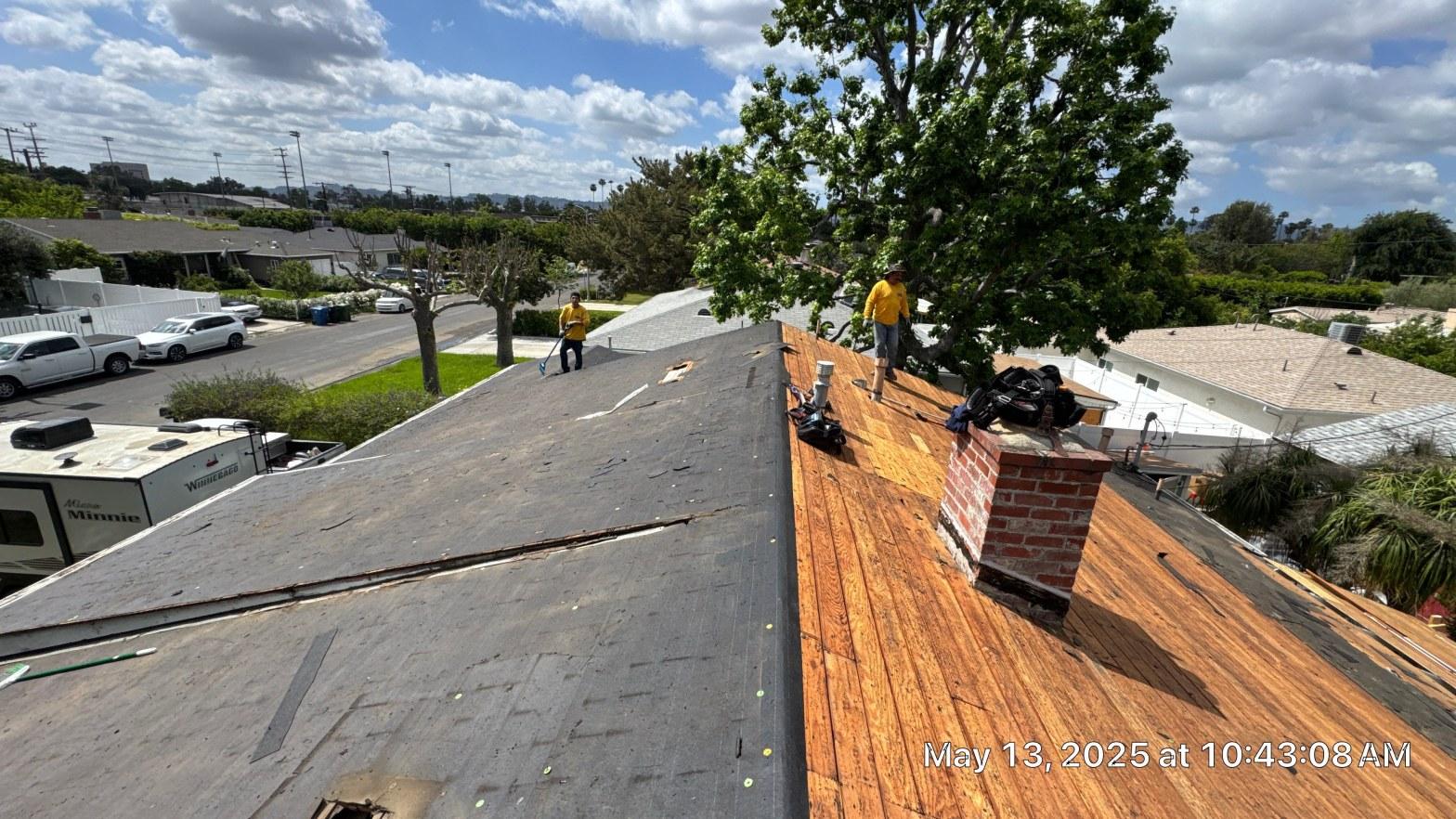 shingle-reroof-6643-saloma-ave-los-angeles-665 Two workers in yellow shirts stripping asphalt shingles from residential roof, exposing plywood decking and brick chimney.