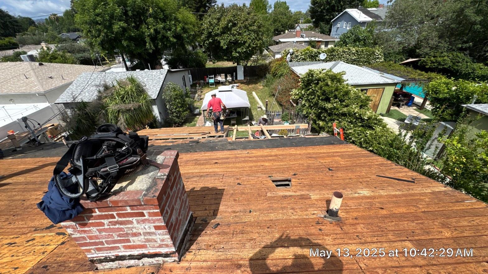 shingle-reroof-6643-saloma-ave-los-angeles-666 Residential roof tear-off showing bare wood decking, brick chimney, pipe penetration, and workers on edge of roof.