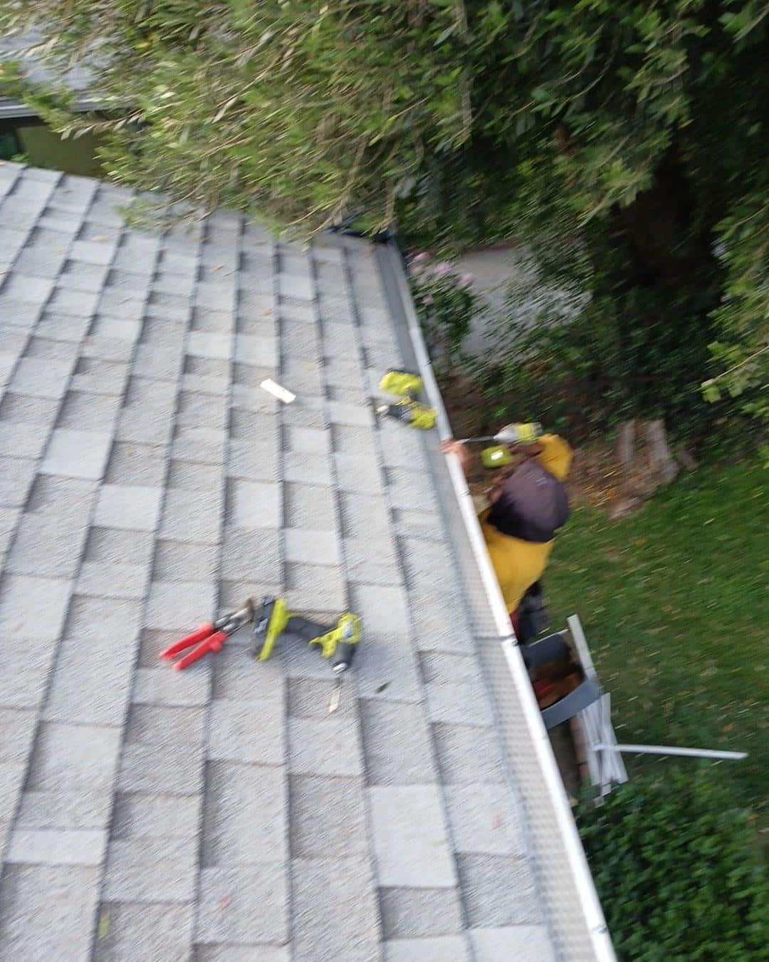 shingle-reroof-6643-saloma-ave-los-angeles-678 Contractor in yellow jacket installing aluminum gutters on asphalt shingle roof, Ryobi tools visible on rooftop.