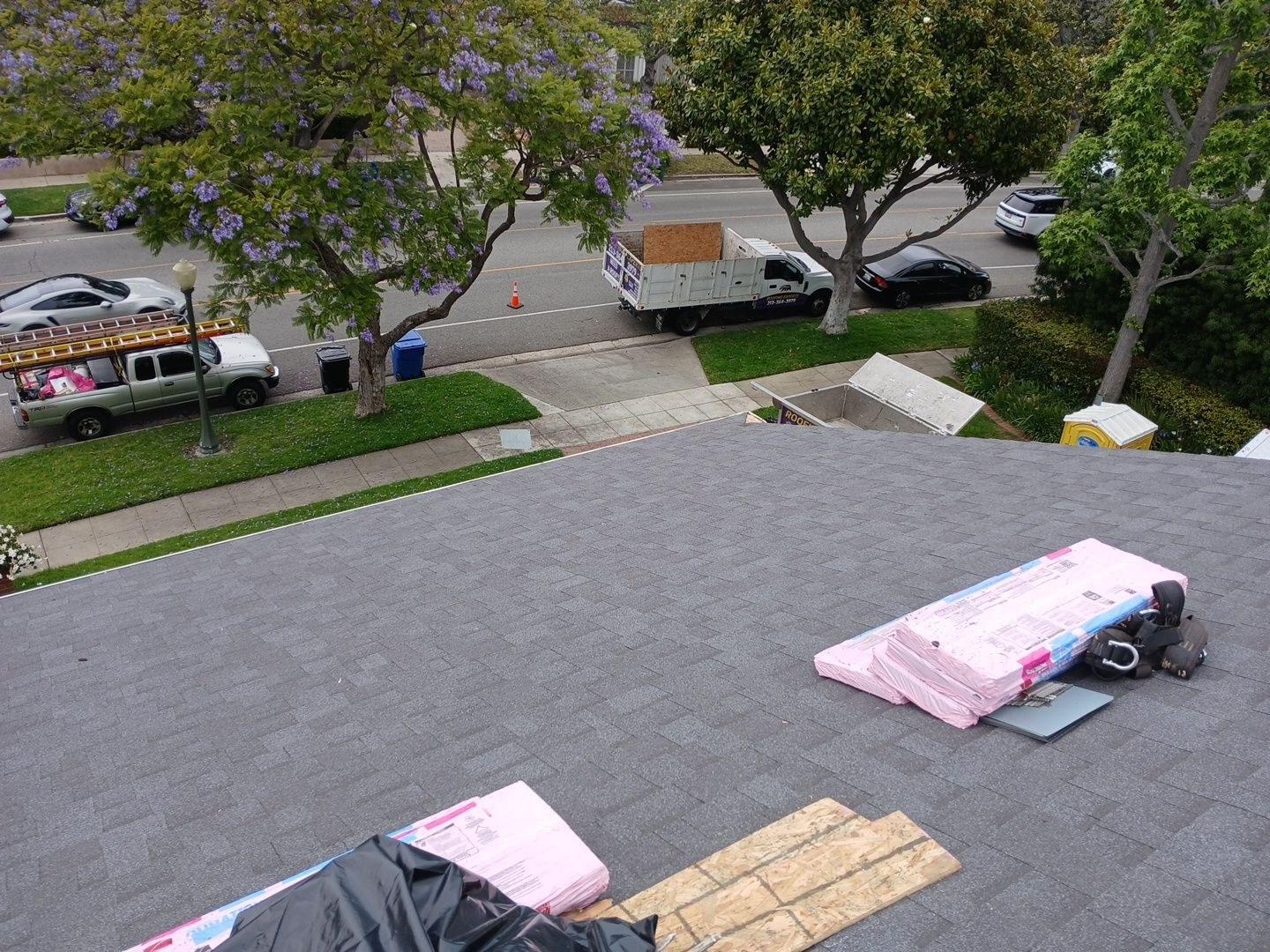 shingle-reroof-motor-ave-los-angeles-603 Freshly shingled gray asphalt roof with pink shingle bundles staged on top, roofing trucks parked on street below.