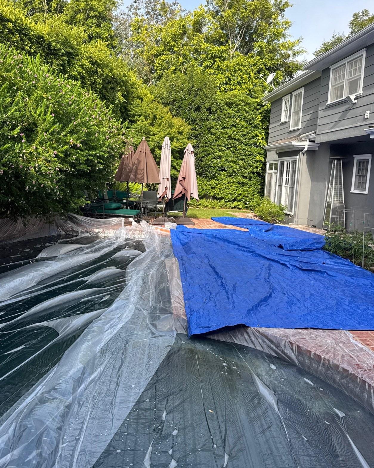 shingle-reroof-motor-ave-los-angeles-610 Residential swimming pool covered with clear plastic sheeting and blue tarp next to gray two-story house and patio umbrellas.